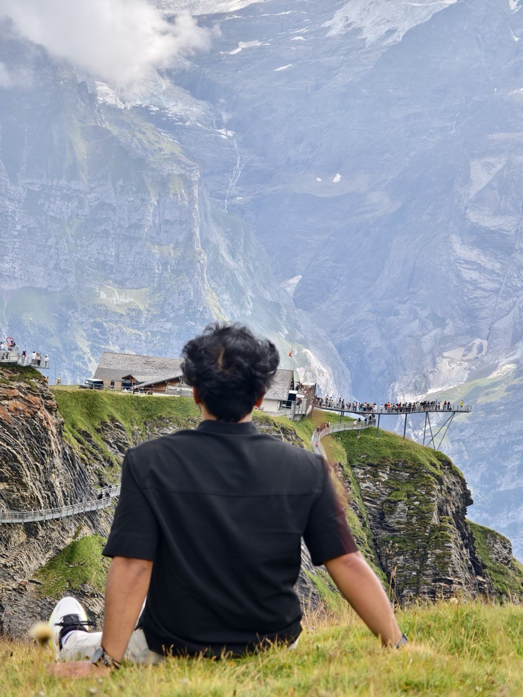 Shivam sitting on the grass at Grindelwald First, looking out at the Cliff Walk and the Alps